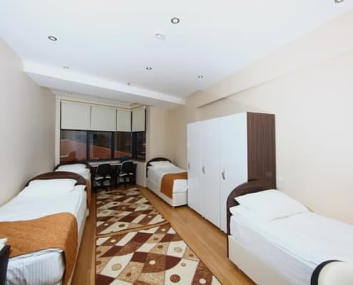 dort-4-kisilik-yurt-odasi dört kişilik yurt odasının giriş kapısı yönünden çekilen fotoğrafı. solda ve sağda ikişer adet yatak, sağda eşya dolapları, karşıda çalışma masaları ve pencere