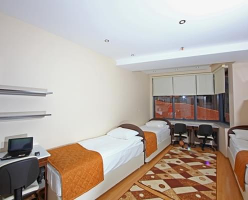 dort-kisilik-kiz-ogrenci-yurdu-odasi Dört kişilik yurt odası giriş kapısı arkaya alınarak çekilmiş fotoğrafı. solda iki yatak, sağda bir yatak, solda çalışma masası ve raflar, karşıda stor perdeyle yarım kapatılımış pencere