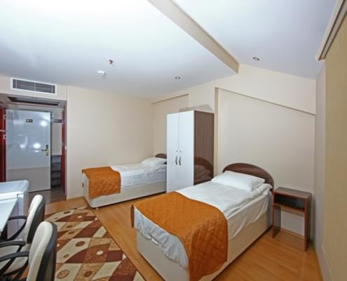 iki-kisilik-oda-aci-2 İki kişilik yurt odasının pencere arkaya alınarak çekilen görseli. solda çalışma masaları ve buzdolabı. Sol ileride giriş kapısı sağda iki adet yatak ve eşya dolabı.