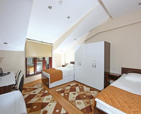 kiz-ogrenci-yurdu-odasi İki kişilik kız öğrenci yurdu odasının giriş kapısı tarafından çekilen fotoğrafı. Saşda iki yatk ve iki kişi için eşya dolanı. Solda üzerinde bilgisayar duran çalışma masası ve karşıda perde ile bir kısmı kapatılmış pencere