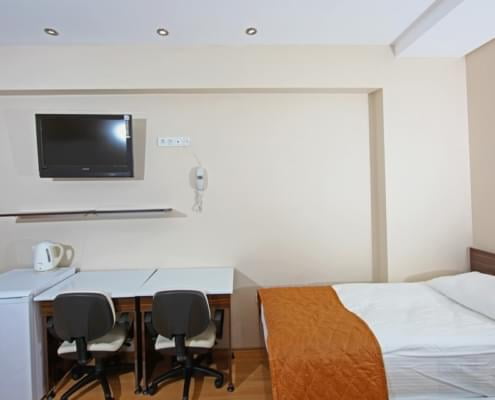 kiz-ogrenci-yurdu-odasi-donanimi kız öğrenci yurdunda bulunan donanımı gösteren fotoğraf. karşıda televiyzon, raf, buzdolabı, çalışma masası. sağda yatak ve stor perde ile kapatılmış pencere.