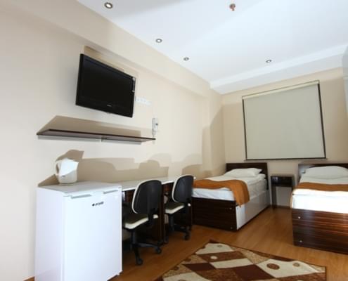 kiz-ogrenci-yurdu-odasi-televizyon iki kişilik kız yurdu odasının giriş kapısı yönünden çekilmiş fotoğrafı. Solda buzdolabı, raf, televizyon çalışma masası ve bir yatak. Sağda bir yatak. Yerde halı.